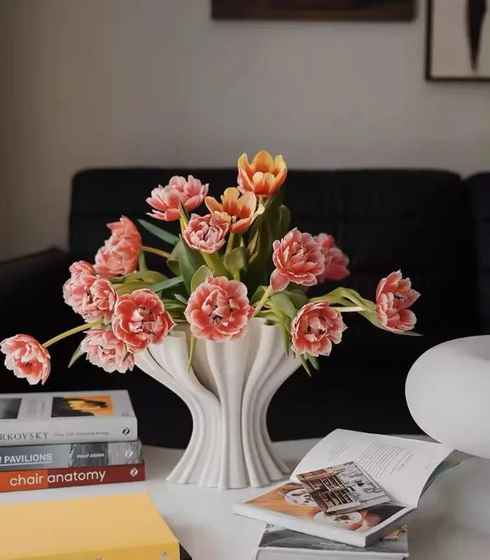 White vase with pink tulips on a table with books and a lamp.
